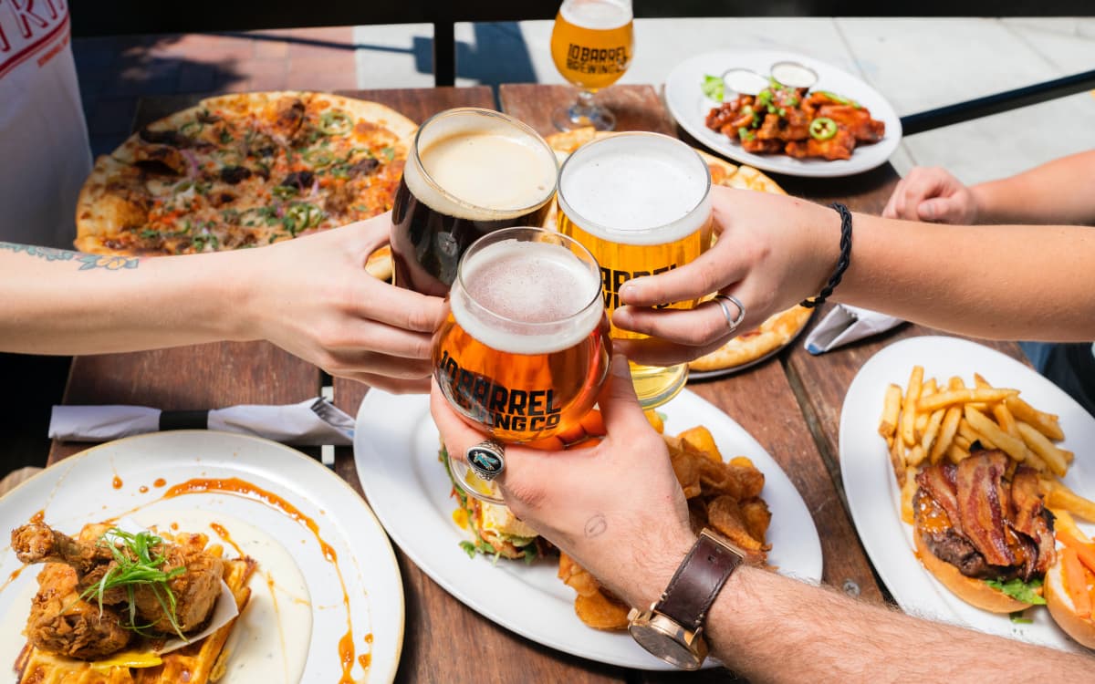 A group of 3 people clicking pint glasses of zero beers together over a table of food